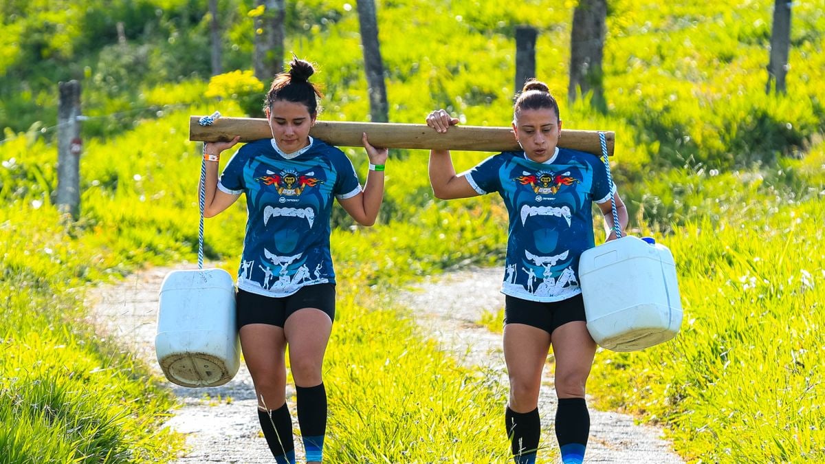 Two women exercise carrying jugs outdoors in a sunny rural area.
